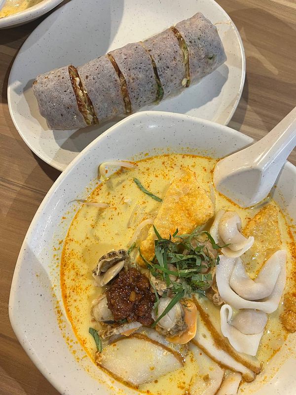 A bowl of laksa accompanied by a roll of rice paper filled with ingredients.