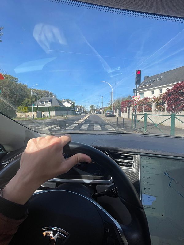 A white Tesla Model S parked at a sunny street with a person walking near it and buildings in the background.