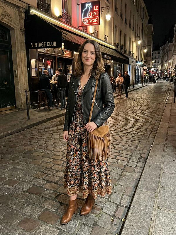 A woman stands on a cobblestone street in Paris at night, dressed in a floral dress and leather jacket.