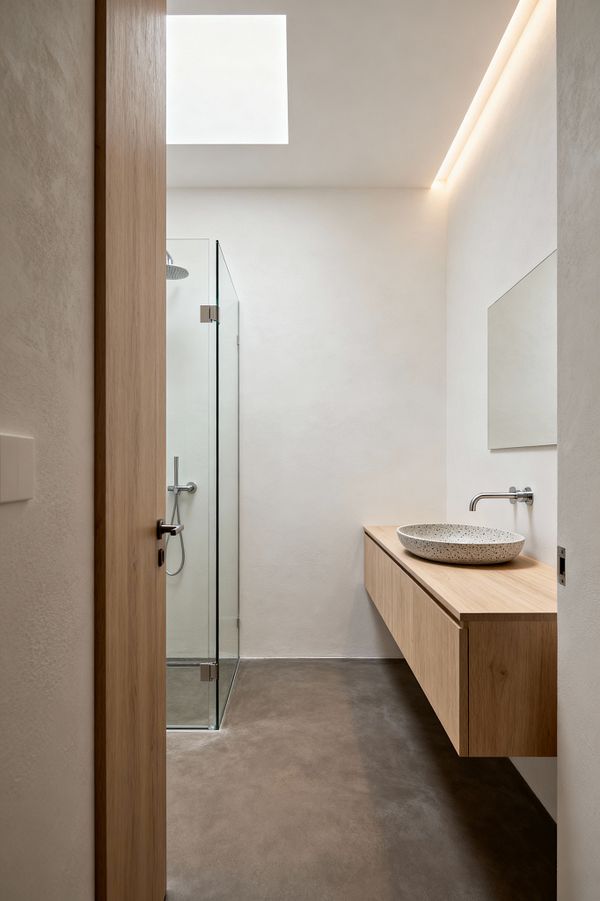 A narrow minimalist bathroom with warm white plaster walls and a large ceiling skylight, softened by concealed cove lighting. To the right, a floating light-oak vanity holds a speckled terrazzo vessel sink with a wall-mounted stainless steel faucet beneath a frameless mirror. At the end of the room, a frameless glass shower with a rain head sits on a smooth gray microcement floor, emphasizing clean lines and open space.