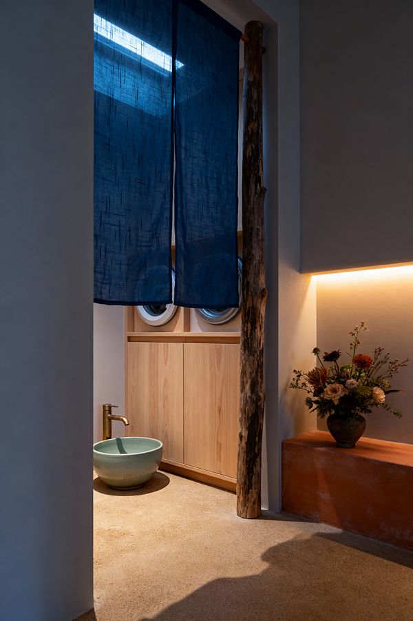 A serene Japanese-inspired laundry room features an indigo linen noren curtain framing a light wood cabinet that conceals front-loading machines, a pale celadon bowl basin with a brass spout, a raw tree trunk post, and an earthen sand-colored floor. To the right, a terracotta plaster ledge holds a simple flower arrangement under warm cove lighting, while a skylight above brings cool daylight across smooth plaster walls.