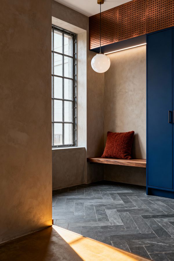 Contemporary mudroom corner with warm taupe plaster walls and a large black steel-framed window. A floating walnut bench holds a rust-colored textured cushion above a charcoal herringbone stone-look floor; to the right, a tall matte navy cabinet with slim black pull sits below perforated copper-toned panels with an integrated linear LED, while an opal-glass globe pendant with a brass canopy hangs overhead and daylight rakes across the floor.