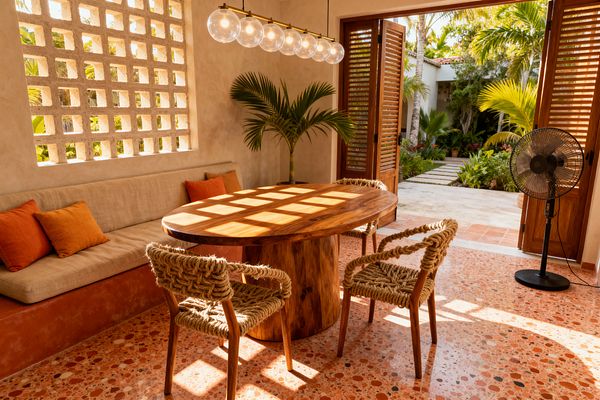 A sunlit tropical dining room with an oval solid wood pedestal table, three rope-woven chairs, and a built-in bench upholstered in beige with burnt-orange pillows. A concrete breeze-block wall filters light across the terrazzo floor in terracotta tones. Louvered wooden doors open to a lush garden, a potted palm sits in the corner, and a linear brass pendant with six clear glass globes hangs overhead; a black pedestal fan stands near the doorway.