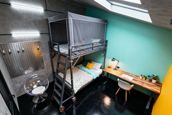 Industrial-style kids' room featuring a steel loft bunk on red casters with a ladder and a gray mesh canopy. The lower bed has white bedding with multicolored dots, a turquoise quilt, and a yellow cushion. To the right, a long butcher-block desk with a black task lamp, pencils, and a small plant sits against an aqua wall with a white chair. The left wall has a concrete-effect finish, linear LED strips, and a perforated metal pegboard. A glossy black floor and skylight complete the space.