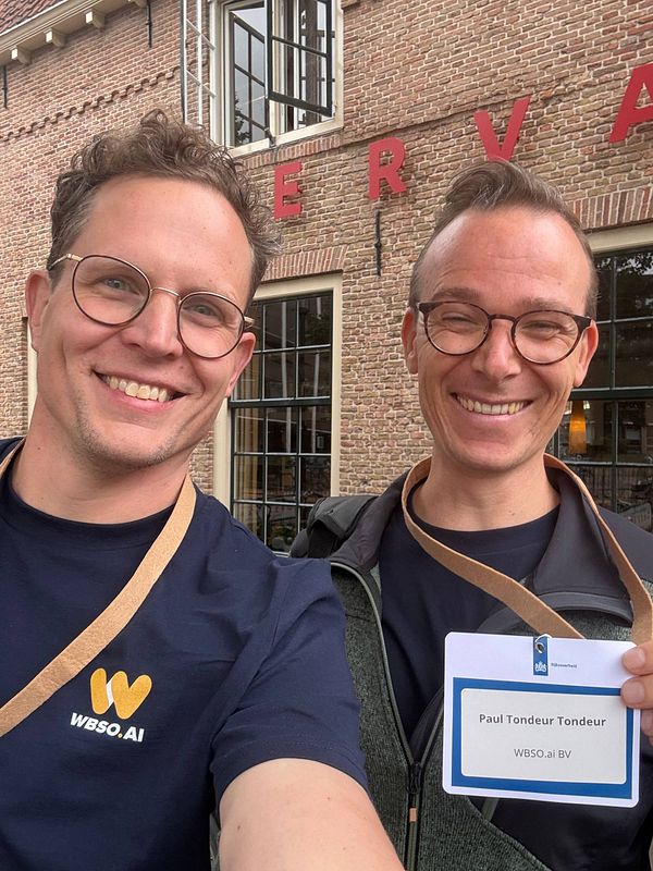 Two smiling men wearing glasses and WBSO AI event badges pose in front of a brick building with large windows and red letters on the wall.