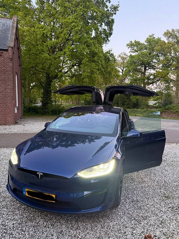 Photograph of a black Tesla Model X parked on a city street beside a sidewalk with a building in the background.
