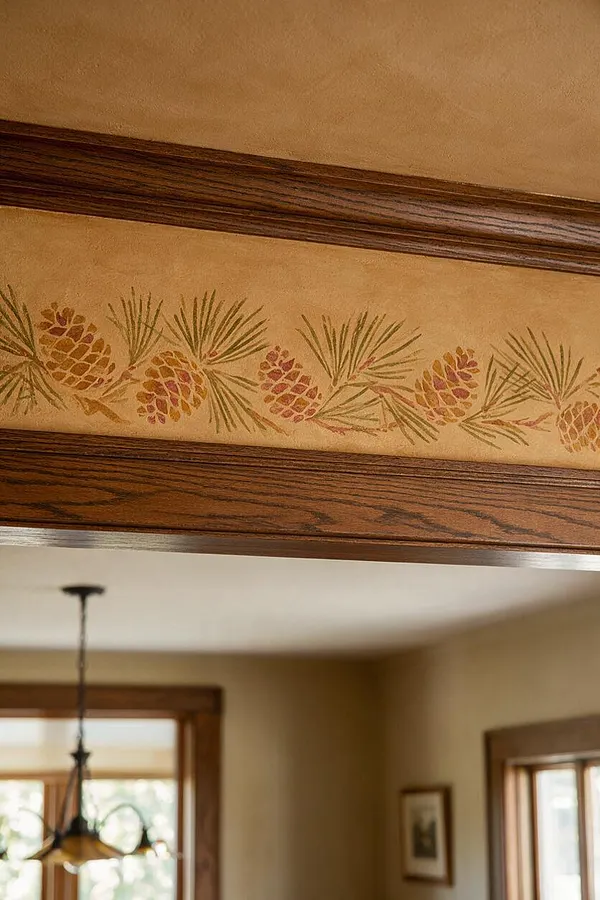 A close-up of a Craftsman-style interior detail showing a tan textured wall band painted with a repeating pinecone and pine needle motif, bordered above and below by rich dark wood trim. Softly blurred in the background are warm beige walls, wood-trimmed windows, and a hanging pendant light, reinforcing the handcrafted, nature-inspired aesthetic.