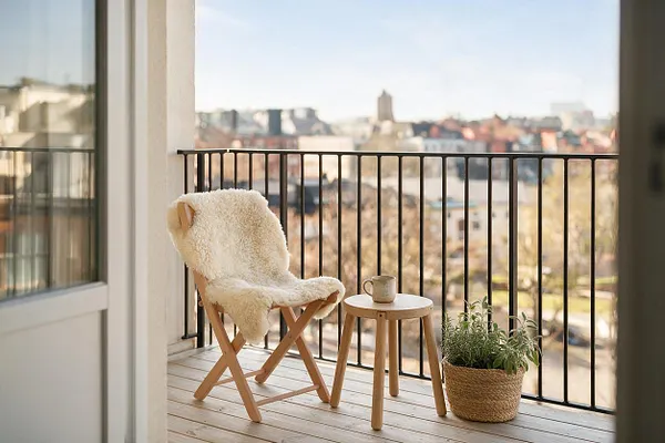 A Scandinavian-style balcony with a light wood folding chair covered in a cream sheepskin throw, a small round wooden side table with a mug, and a woven basket planter filled with greenery. Black metal railings frame a soft, sunlit city view in the background.