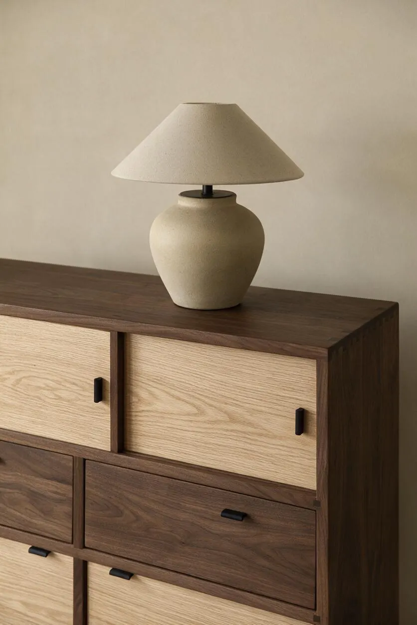 A minimalist Japandi-style table lamp sits on a wooden dresser with mixed light oak and dark walnut drawer fronts. The lamp has a cream ceramic base and a tapered beige shade, complementing the warm neutral wall behind it.