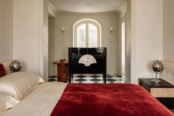 A master bedroom in Art Deco style with a bed dressed in cream bedding and a deep red velvet throw in the foreground. At the back of the room, a glossy black cabinet with a shell motif stands on a black-and-white checkered floor beneath an arched window, flanked by wall sconces and framed by soft neutral walls.