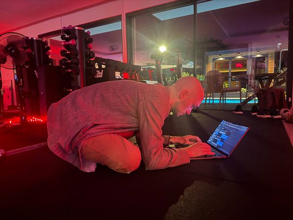 Person working on a laptop while kneeling on the floor in a dimly lit room with red lighting and gym equipment.