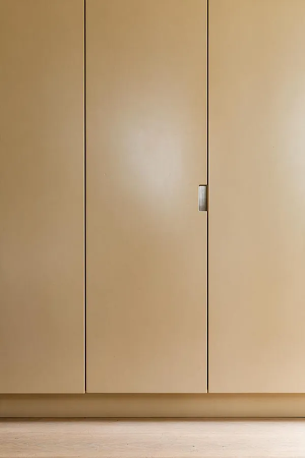 A close-up view of flat-panel beige cabinet doors in a minimalist interior, with slim vertical seams and a small recessed metal handle on one door. The cabinets sit above a light wood floor, creating a calm neutral palette.