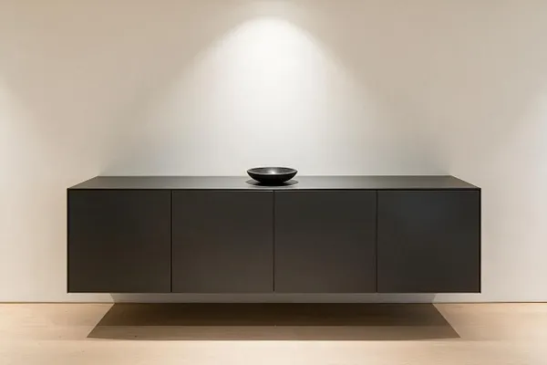 A minimalist interior vignette with a floating dark charcoal cabinet mounted against a warm white wall, centered beneath a soft spotlight. A small black bowl sits on top, and the pale wood floor and subtle shadowing emphasize the clean, sculptural composition.