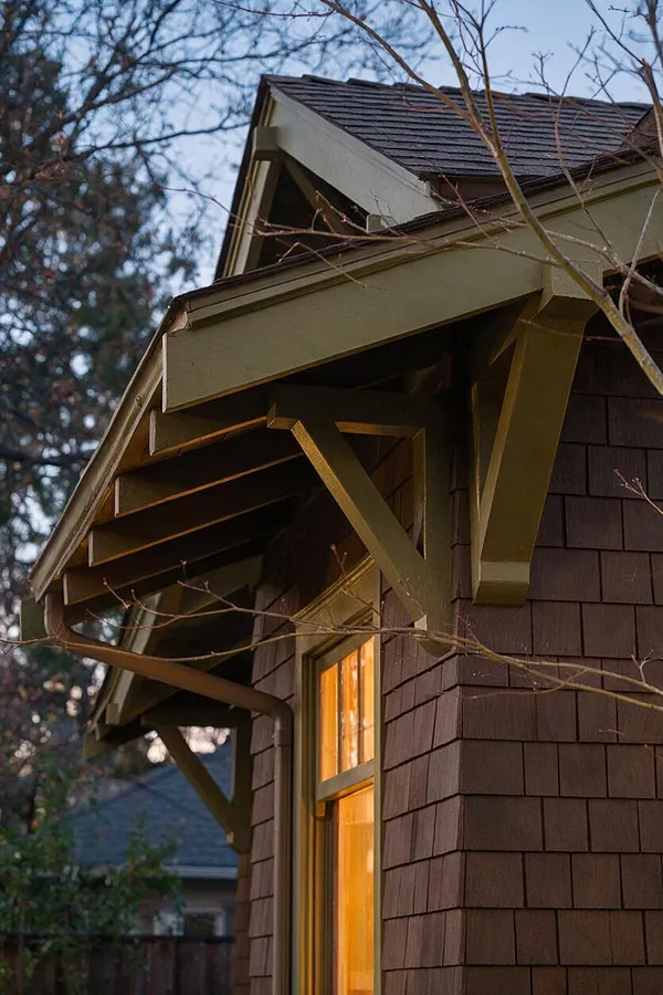 A close-up of a Craftsman-style house exterior showing a steep gable roof, exposed triangular rafter brackets, and wide olive-green trim against dark brown shingle siding. A single window glows with warm interior light beneath leafless tree branches, with dusk sky and neighboring trees in the background.