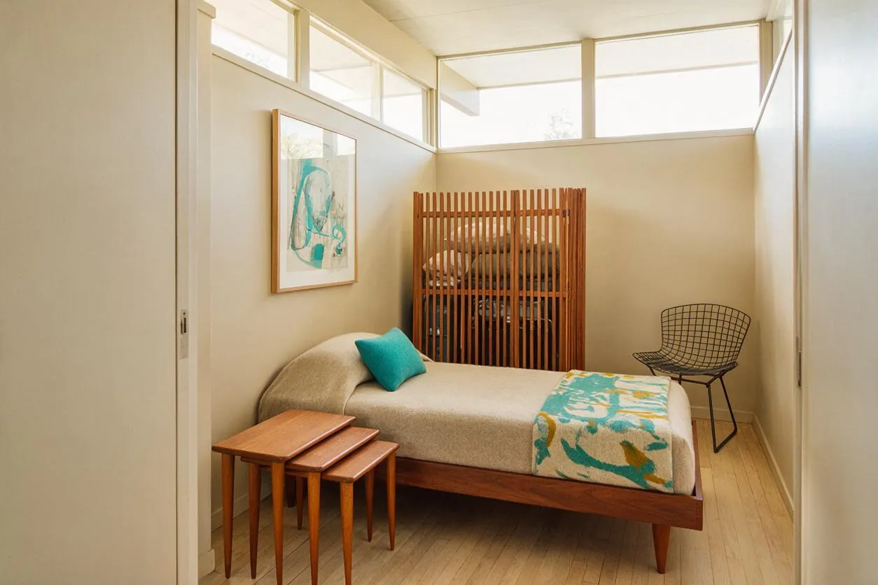 A compact guest bedroom with a low wooden bed frame, beige bedding, and a turquoise throw and pillow. The room includes a wire accent chair, nested wooden side tables, a framed abstract artwork, and a slatted wood screen, all under high clerestory windows that fill the space with soft daylight.
