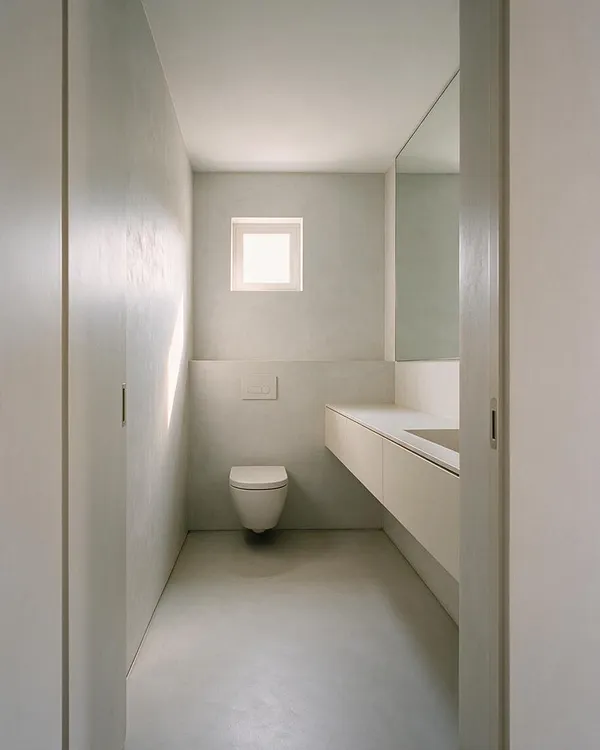 A narrow minimalist powder room with pale gray walls, a small square frosted window at the back, and a wall-hung toilet centered below it. On the right, a long floating vanity with a white counter and integrated sink extends toward the camera, while a large mirror above reflects light and enlarges the compact space.