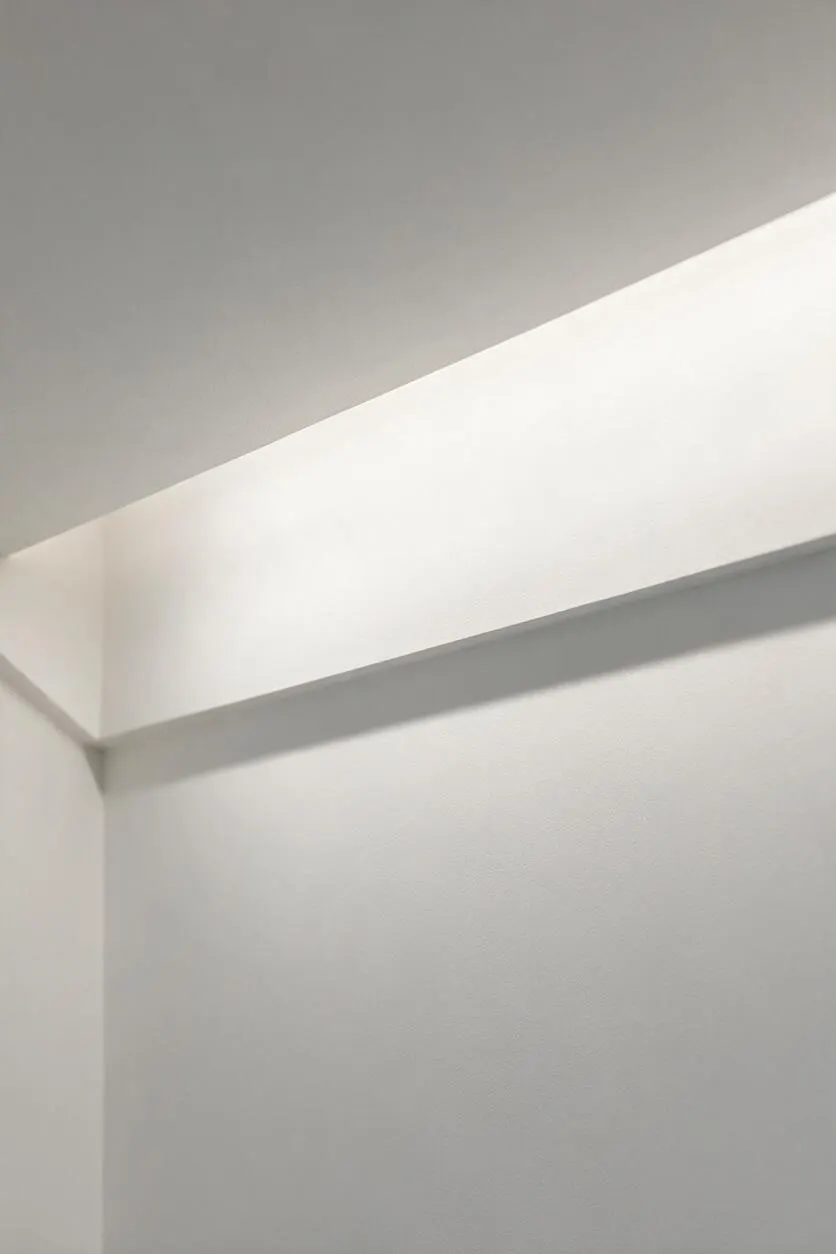 A close-up of a minimalist interior wall and ceiling corner painted in soft white and light gray. A clean horizontal architectural ledge casts a subtle shadow across the smooth surface, creating a simple geometric composition with no decoration.