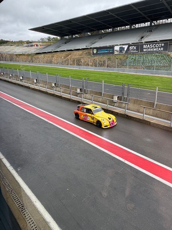 A small colorful race car on the pit lane at Circuit Spa-Francorchamps with empty grandstands in the background.