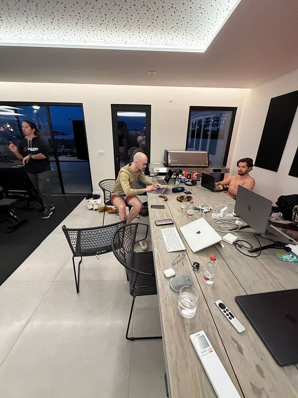 A minimalist indoor workspace setup featuring a wooden table with two laptops and a dual-monitor desktop computer, accompanied by office furniture and plants.