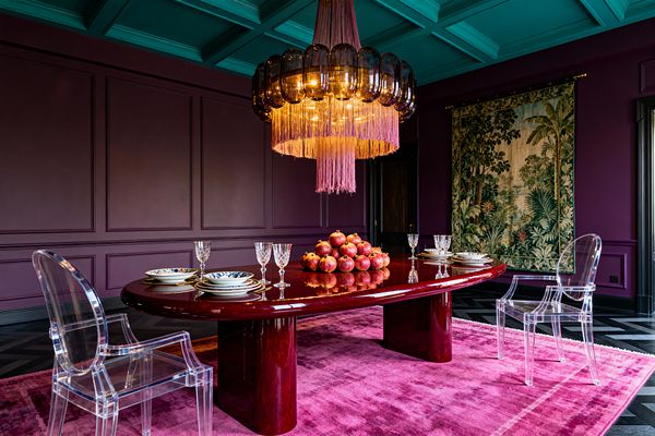 A dramatic dining room with deep plum paneled walls and a teal coffered ceiling features a large oval burgundy lacquer table set with crystal stemware and blue-and-white china. Clear acrylic armchairs sit on a saturated pink rug over a black chevron wood floor. Overhead, a chandelier of smoked glass and warm bulbs is finished with mauve fringe. A verdant landscape tapestry hangs on the right wall; a mound of pomegranates forms the centerpiece.