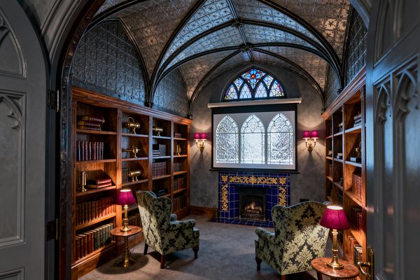 A compact Victorian-style media room with a groin-vaulted pressed-tin ceiling and dark ribs, lined by walnut bookcases filled with leather-bound books and brass objects. Two green damask wingback chairs face a drop-down projector screen above a cobalt blue, gold-embellished tile fireplace, with charcoal gray walls, stained-glass tracery, and brass sconces with plum shades casting warm light.