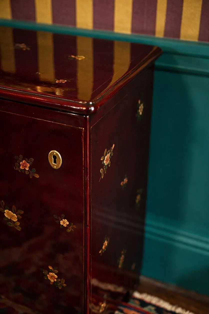 A close-up of a glossy burgundy lacquer cabinet decorated with tiny painted floral motifs and a brass keyhole plate. It sits against a teal wall with gold-and-plum vertical striped wallpaper, creating a rich maximalist contrast.