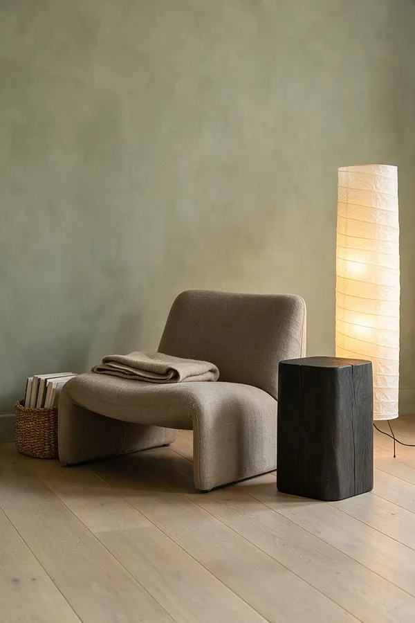 A minimalist Japandi-style corner with a taupe upholstered lounge chair, a folded beige throw, a woven basket of books, a tall white paper floor lamp, and a dark wooden side table on pale wood flooring. The soft green wall and warm lighting create a calm, earthy atmosphere.