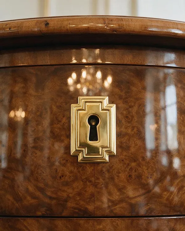 A close-up of a polished burl wood cabinet front with a glossy curved top edge and a centered geometric brass keyhole escutcheon. The warm brown wood grain is highly reflective, with soft blurred light reflections in the surface, emphasizing the elegant Art Deco detailing.