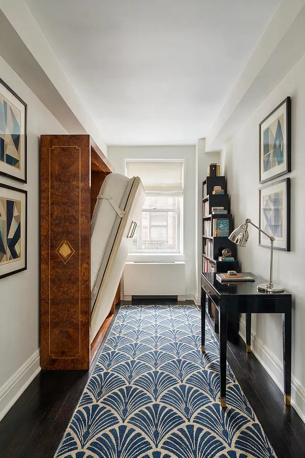 A narrow studio apartment interior with a fold-down bed concealed inside a glossy burl wood cabinet on the left, a black writing desk and shelving on the right, and a blue-and-cream geometric rug running down the center. Framed abstract prints line the walls, while a bright window at the end of the room adds natural light.