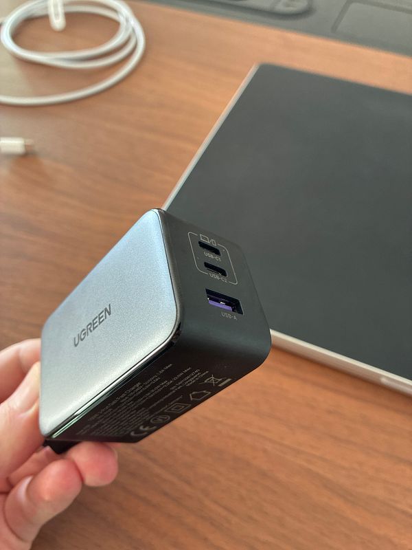Close-up of a UGREEN 100W GaN charger held by a hand, with a laptop and charging cable on a wooden desk background.
