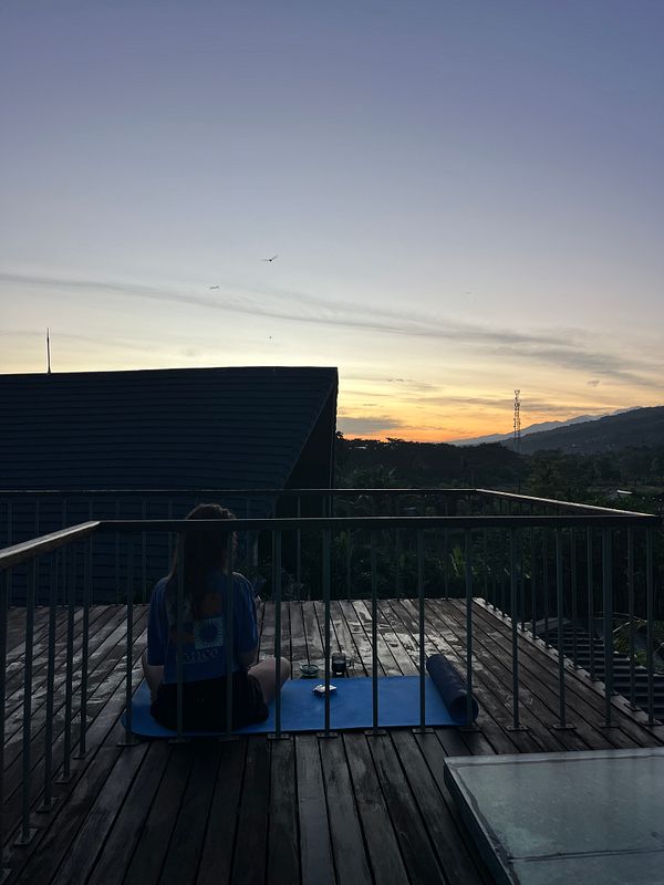 A person sits on a yoga mat on a balcony, gazing at a colorful sunrise over the horizon.