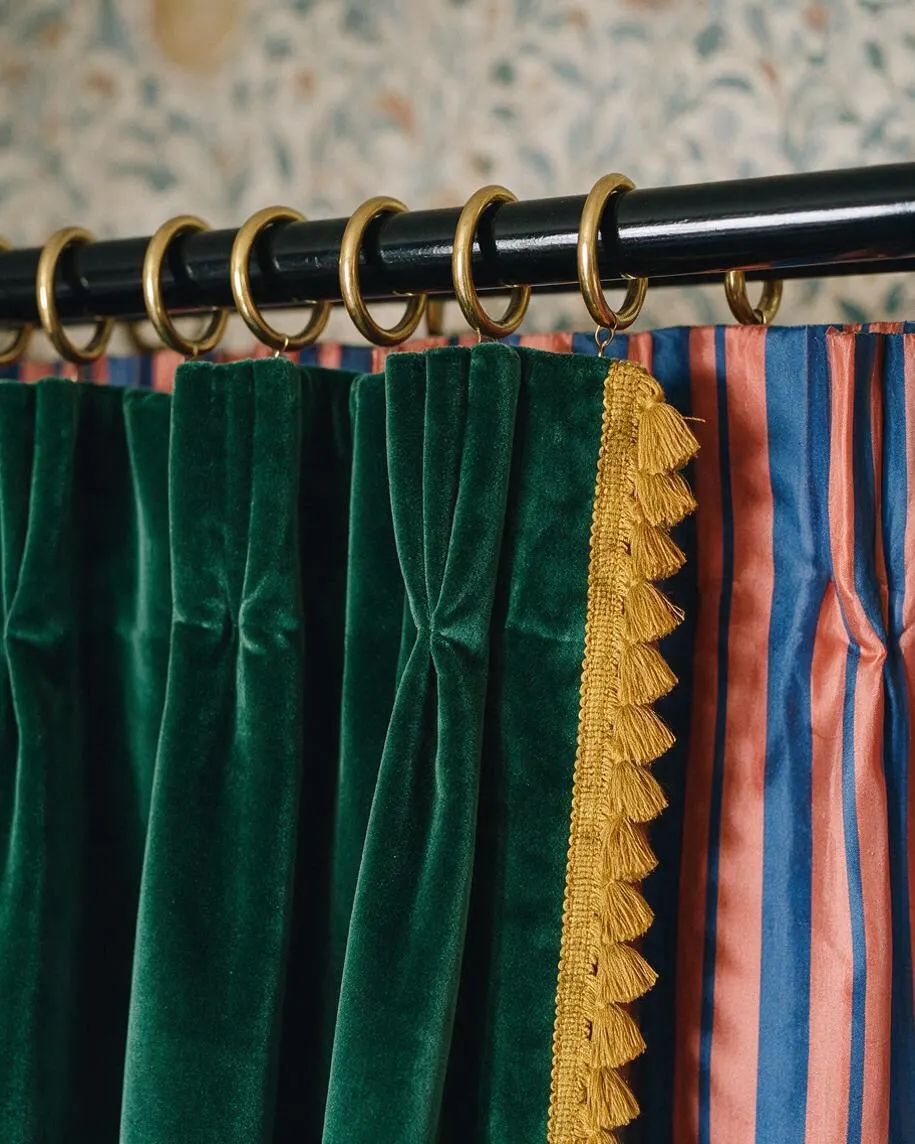 A close-up of layered curtains hanging from a black rod with brass rings. Deep emerald velvet panels are paired with striped pink and blue fabric, finished with a gold tassel trim, creating a rich, maximalist look.