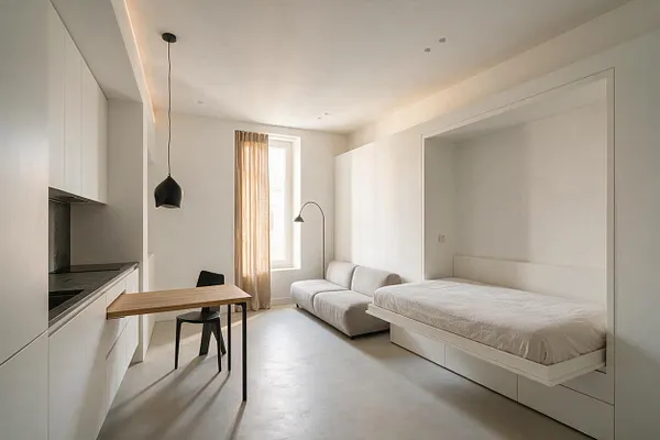 A bright minimalist studio apartment with white built-in cabinets and a compact kitchen on the left, a small wood dining table with a black chair in the center, and a low bed tucked into a recessed alcove on the right. A light gray sofa, black pendant light, and sheer curtains at the window soften the clean, neutral space.