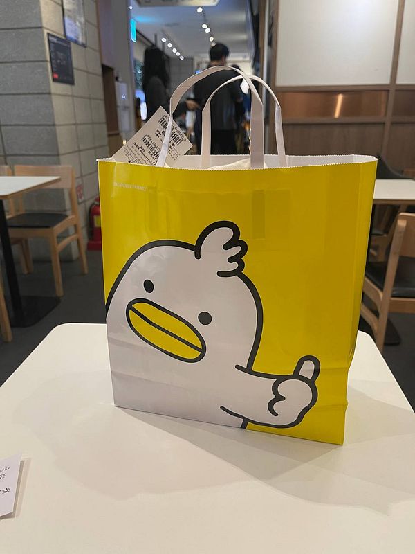 A bright yellow shopping bag featuring a cartoon duck design is placed on a table in a cafe.
