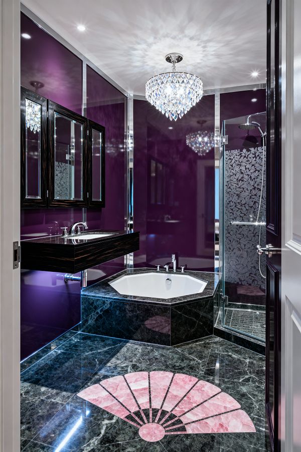 A glamorous Art Deco bathroom features high-gloss plum wall panels framed in chrome, a faceted crystal chandelier, and black marble flooring with a pink fan inlay. An octagonal white soaking tub is set into a black marble base beside a floating dark-wood vanity with a polished top, while a glass shower enclosure shows an etched floral panel and chrome rain shower; recessed spots and reflective surfaces amplify light across the compact space.