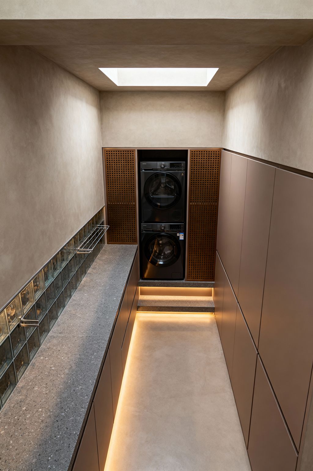 A narrow contemporary laundry room features matte taupe, handleless cabinets on both sides and a gray stone-look countertop along the left wall, backed by a short run of square glass blocks with a slim stainless drying rail. At the far end, a stacked black washer and dryer are recessed between perforated wood panels and accessed by a small step with integrated LED strips; warm beige microcement walls and floor and a central skylight provide soft, even light and a minimalist feel.