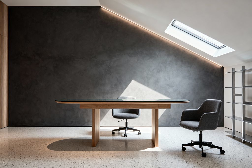 A contemporary home office features a glass-top desk on a light wood base centered on a pale terrazzo floor. Two gray upholstered swivel chairs with black casters sit before a charcoal microcement wall. A sloped ceiling with a skylight and concealed linear LED washes the wall and floor with daylight. To the right stands open stainless-steel shelving against a white partition, while vertical wood slats on the left complete the clean, minimalist space.