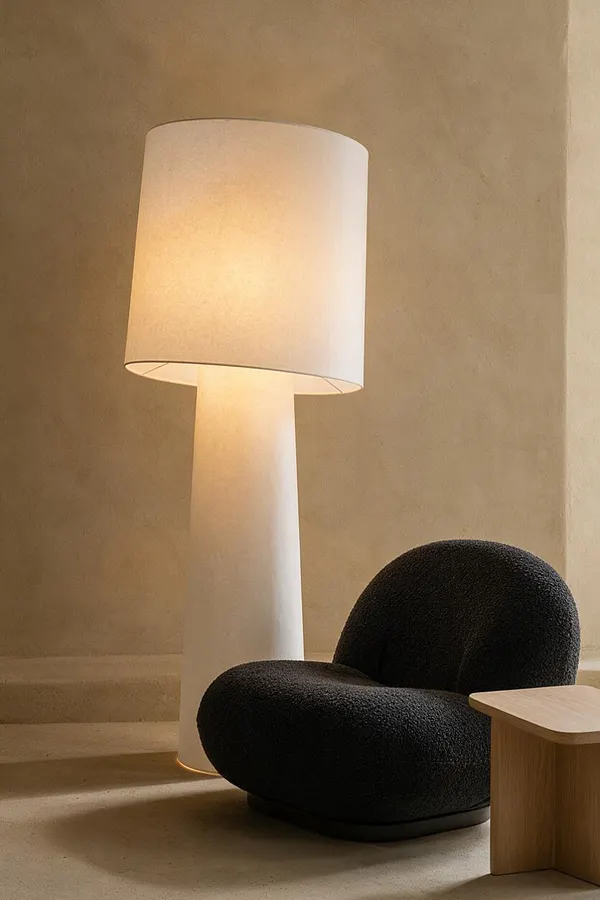 A serene Japandi-style corner featuring a tall white floor lamp with a cylindrical shade, a sculptural black boucle lounge chair, and a small light wood side table. The space uses warm beige walls and a soft neutral floor to create a calm, minimalist atmosphere.