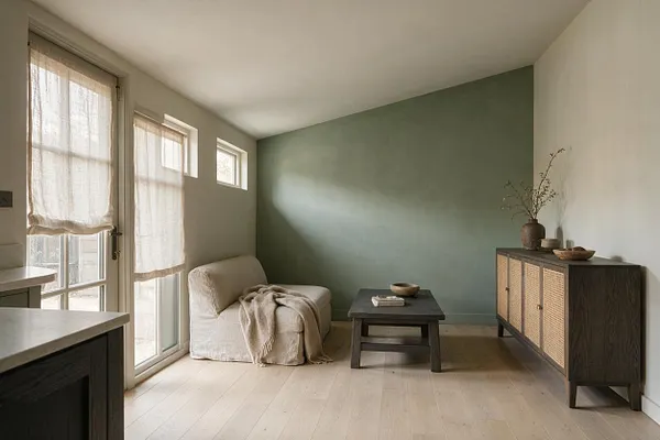 A calm Japandi-style patio with light wood flooring, a sage green accent wall, and soft beige walls. A low cream lounge chair with a draped throw sits beside a dark wood coffee table, while a woven-front cabinet and simple ceramic decor add natural texture and warmth.