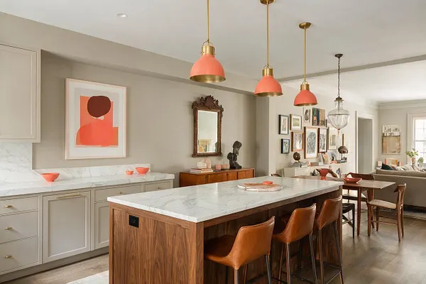 A spacious eclectic kitchen features a large wood island with a white marble countertop and tan leather bar stools. Taupe cabinets, coral pendant lights, framed artwork, a carved mirror, and mixed vintage-style decor create a layered, collected look.