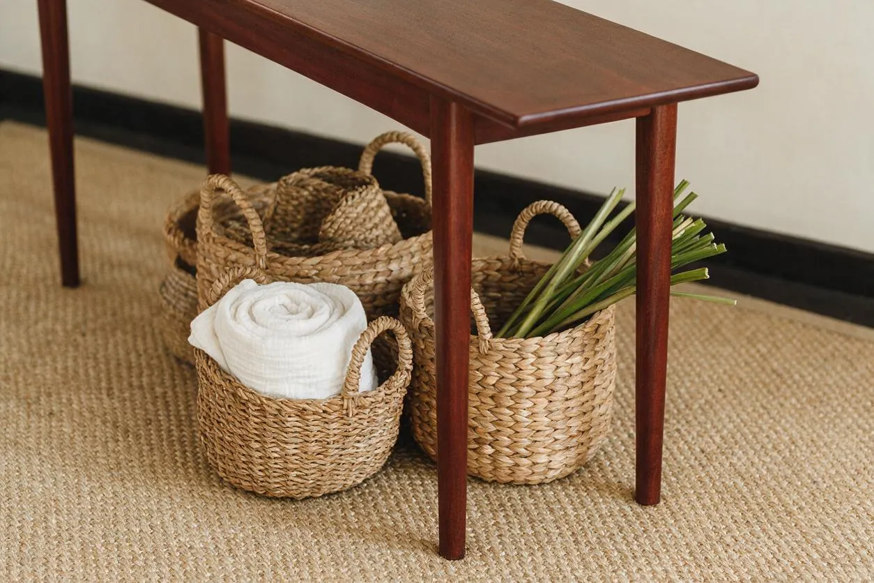 A narrow reddish-brown wooden table stands over several woven natural-fiber baskets arranged on a textured jute rug. One basket holds a folded white towel, another contains green palm-like leaves, and the scene has a warm, relaxed tropical feel.