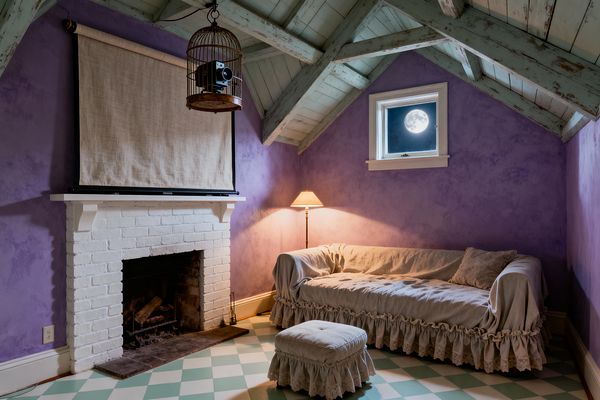 An attic media room with lavender plaster walls and a distressed mint beamed ceiling. A white-painted brick fireplace holds a linen roller screen, while a vintage film projector hangs inside a metal birdcage. A ruffled linen slipcovered sofa and matching ottoman rest on a pale mint-and-cream checkerboard painted floor. A slim floor lamp with a pleated shade glows beside a small window showing the moon, creating a cozy, romantic atmosphere.