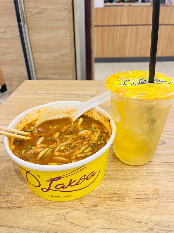 A bowl of Asam Laksa and a drink are placed on a wooden table.