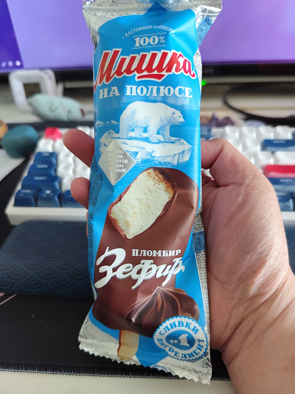 A hand holds a packaged ice cream treat in front of a computer setup.