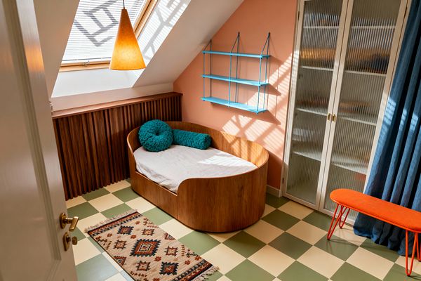 A compact kids' room under a sloped ceiling features a skylight and cone pendant casting patterned light on a peach wall, where bright blue shelves hang above a curved walnut-veneer daybed with neutral bedding and teal round and bolster pillows beside a fluted wood radiator cover. The green-and-cream checkerboard linoleum floor holds a small kilim runner, while a white wardrobe with reeded-glass doors, denim-blue drapery, and an orange hairpin-leg bench complete the mid-century look.