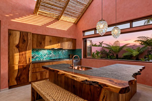 A tropical kitchen with coral plaster walls and a bamboo slatted roof fills with sunset light, with warm hardwood cabinets along one wall above a teal fish-scale tile backsplash. A live-edge wood island with a honed gray stone counter and copper sink sits beneath two capiz shell pendants, while wide windows open to a courtyard of palms and ferns against a matching coral wall.
