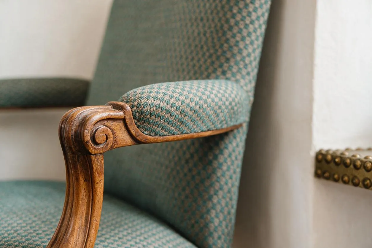 A close-up view of a vintage upholstered armchair with a carved wooden armrest and teal patterned fabric. The chair sits beside a light wall with a small section of brass nailhead trim visible in the background, highlighting the mix of classic detailing and textured fabric.