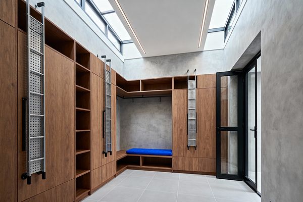 An L-shaped modern mudroom lined with floor-to-ceiling walnut cabinetry includes open cubbies, a central hanging rod, and a bench with a vivid blue cushion. Smooth gray microcement walls and large light-gray porcelain tiles set a neutral base, while clerestory windows and slim recessed ceiling lights wash the space with daylight. A black-framed glass door opens outdoors, and vertical aluminum ladders on black brackets allow access to upper storage.