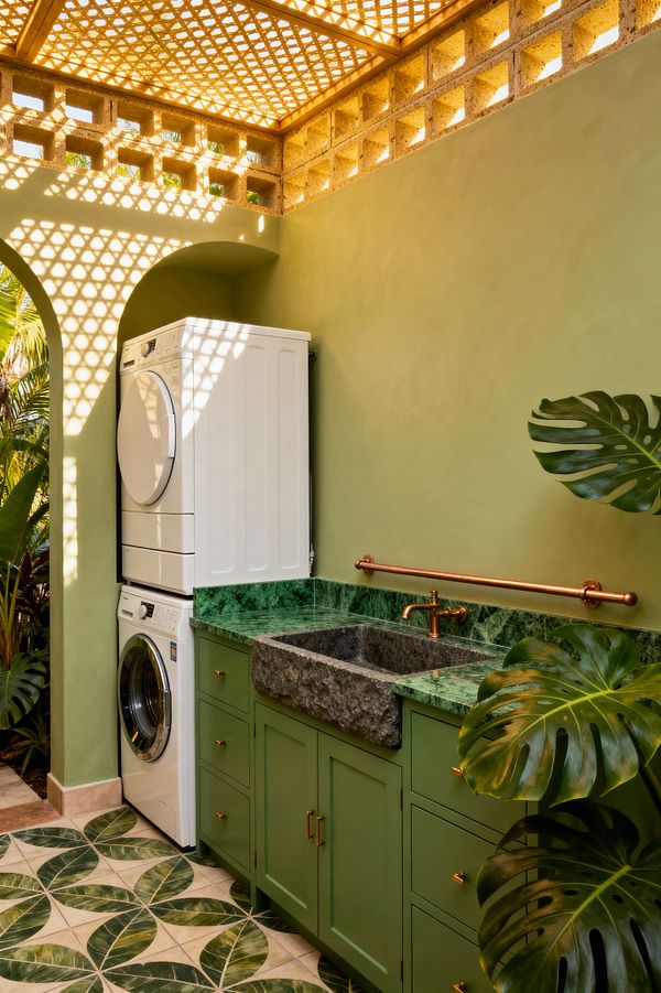 A compact tropical laundry shows a stacked white washer and dryer beside olive-green base cabinets with a deep green marble countertop. A rough-cut stone sink and wall-mounted copper faucet sit against sage plaster walls, while dappled light filters through a wood lattice ceiling and concrete breeze blocks; leaf-pattern ceramic floor tiles and large monstera leaves emphasize the indoor-outdoor feel.