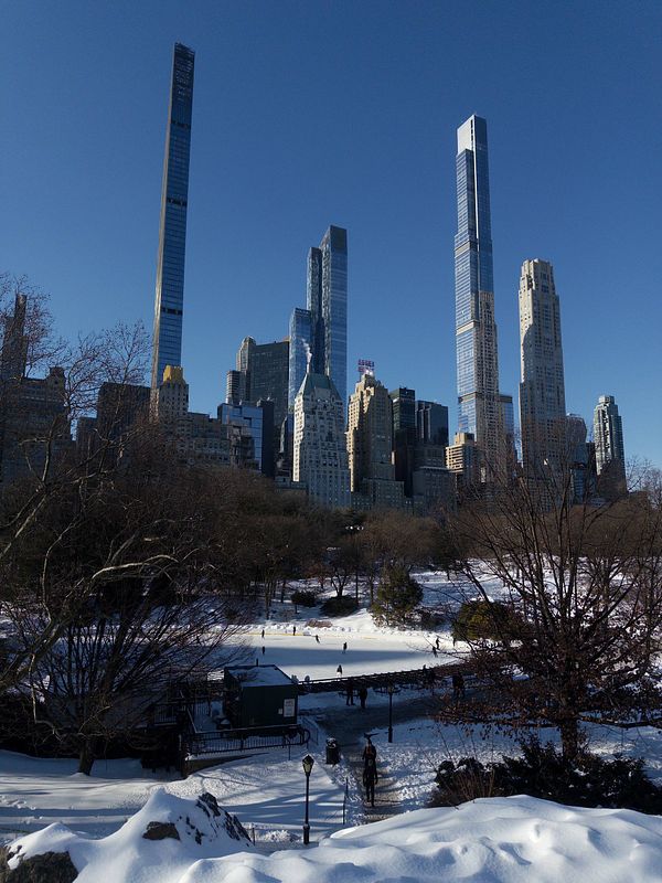 Panoramic aerial view of Central Park surrounded by New York City skyscrapers under a partly cloudy sky.